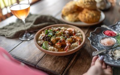 A glass of wine, a dish of shrimp and rice, an oyster platter and a plate of biscuits on a wooden table.